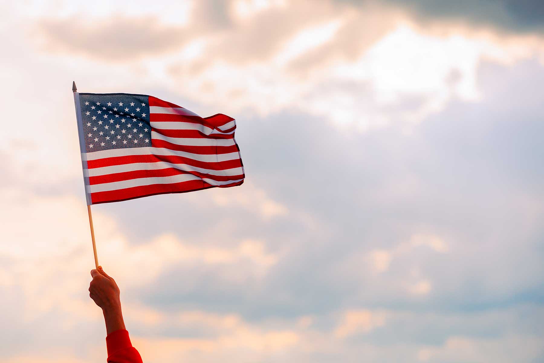 A raised hand waves a miniature American flag proudly under a bright sky.