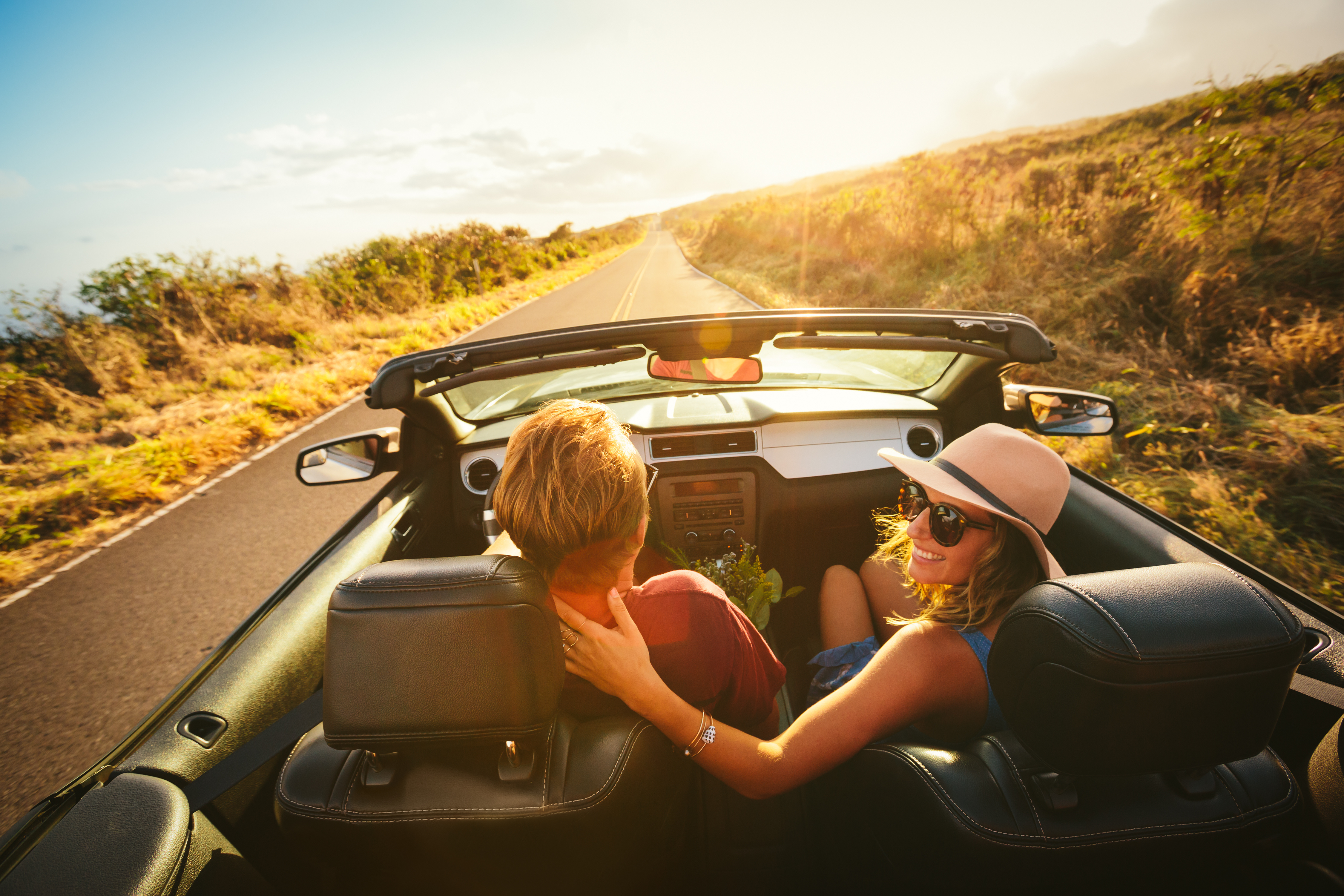 A man and a woman drive along a scenic road in a convertible with the top down, enjoying the open air and sunshine. The woman is smiling, hair blowing in the wind, while the man focuses on the road ahead.