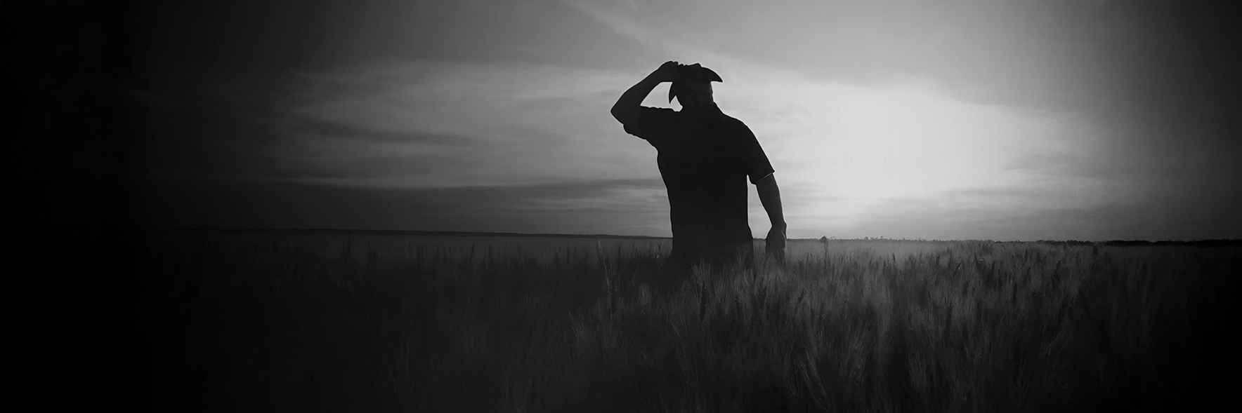 Close-up of a farmer's muddy boots walking through a crop field with a tractor working in the background, captured in black and white.